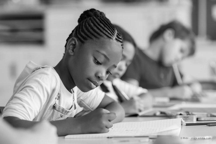An elementary aged girl sits at her classroom desk, writing on a notebook. A boy student is in the background behind her and is also writing in a notebook.