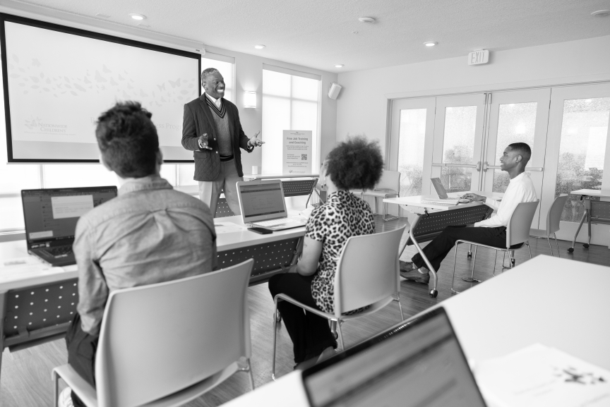A man is presenting to a group of 3+ adults in a meeting room setting. 