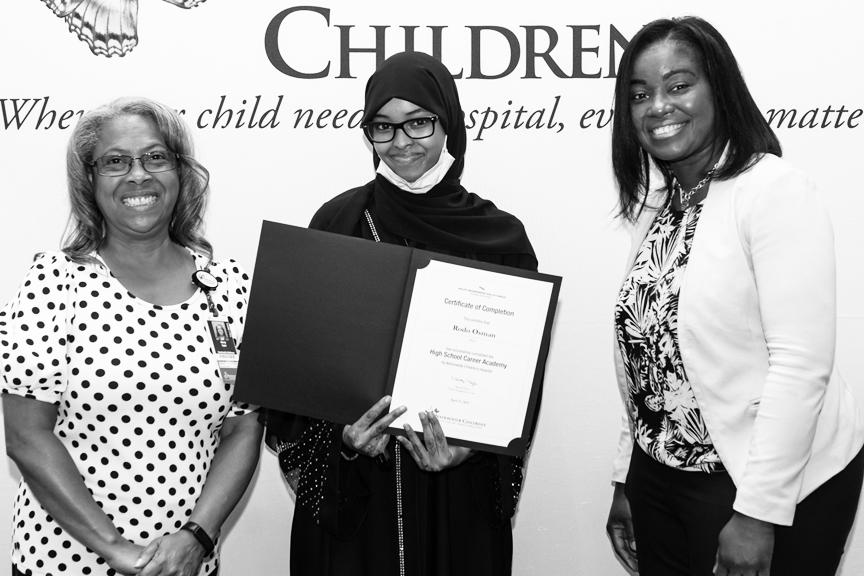 A high school student stands with two employees, holding a certificate of completion