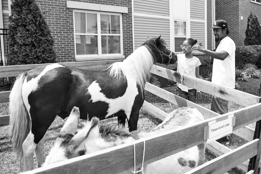 A child and her father pet a pony in an outdoor petting zoo in the neighborhood.