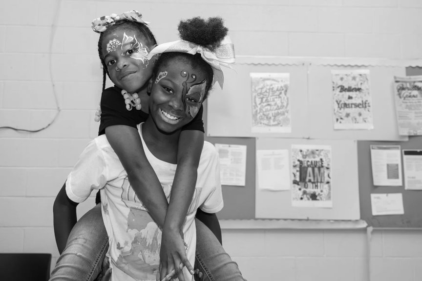 A friend receives a piggy back ride from her friend while playing indoors. Both girls have intricate face paint from the PlayStreets event.