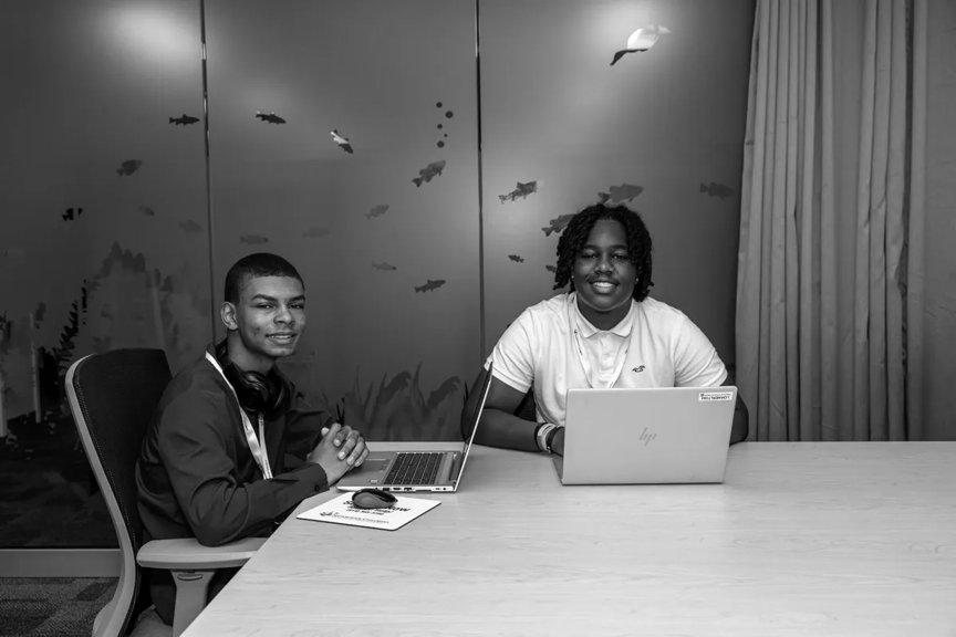 Two high school senior boys are smiling, seated at a conference table. Both have laptops in front of them. 