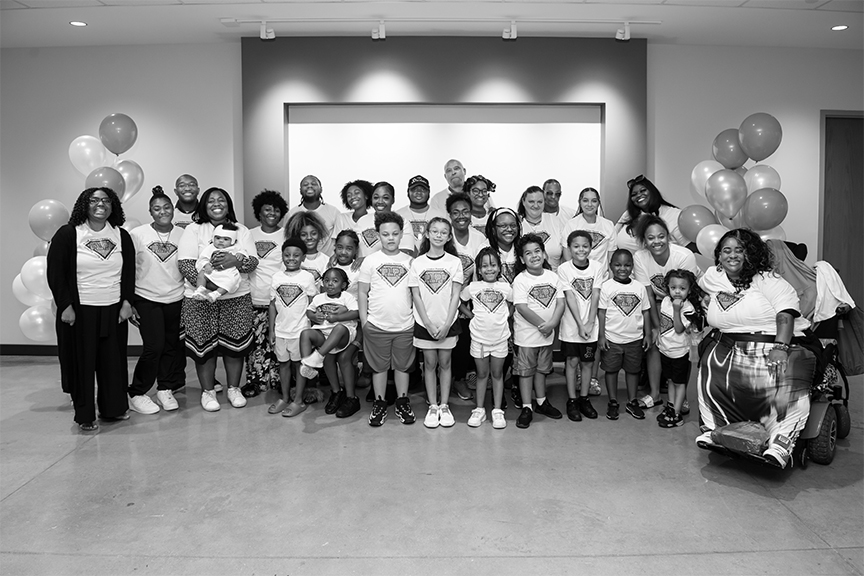Large, smiling group of parents and kids wearing matching shirts. There are balloons in the background.