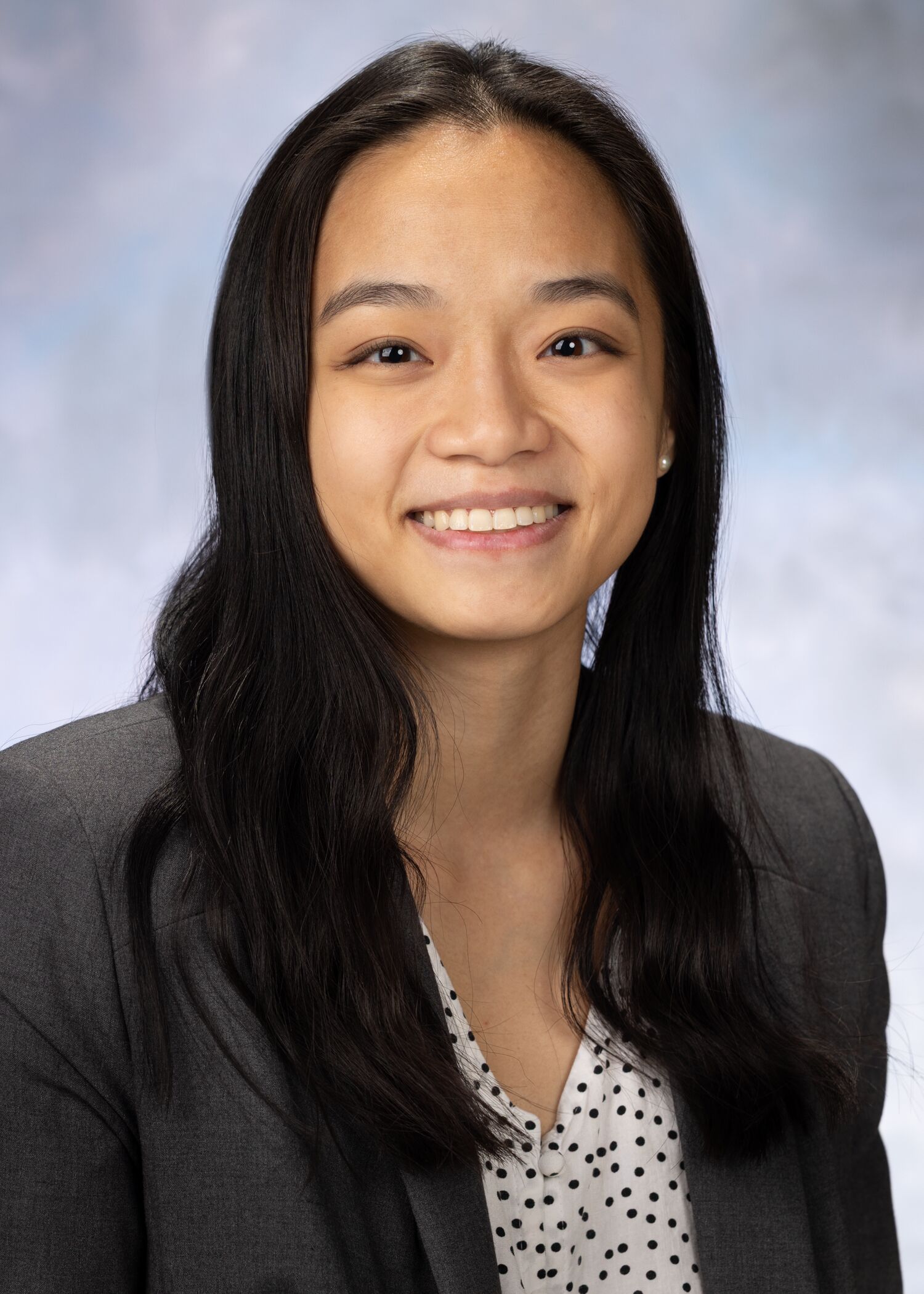 Portrait of Carly Schafer. Carly has long dark hair and is wearing a white and black spotted top with a gray blazer.