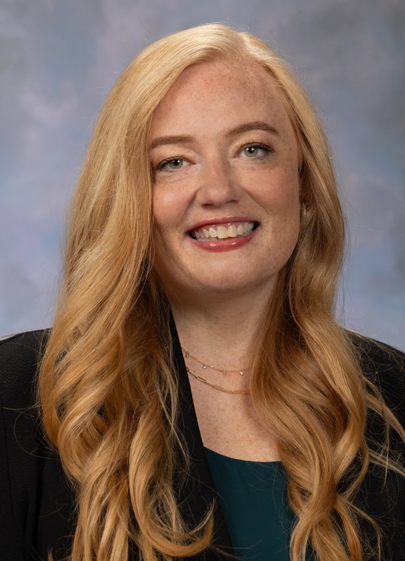 Portrait of Kelly McNally. Kelly has long strawberry blonde hair. She is wearing a dark green top and a black blazer. 