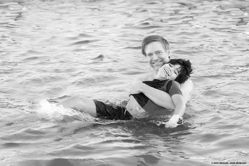A father is holding his sun in a body of water. They are both smiling at the camera.