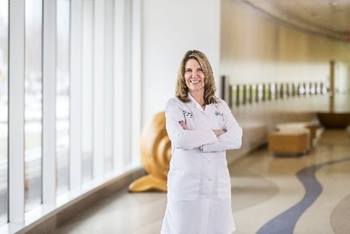 Dr. Catherine Krawczeski stands confidently in a bright hospital hallway, smiling with arms crossed while wearing a white medical coat. Natural light pours in from large windows, creating a welcoming and professional setting.