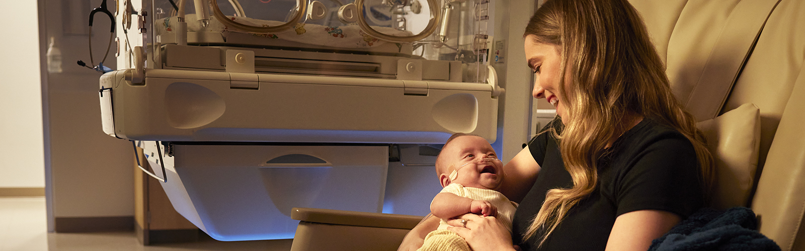 Baby Canaan is smiling, wrapped in his smiling mom's arms, in a NICU setting.