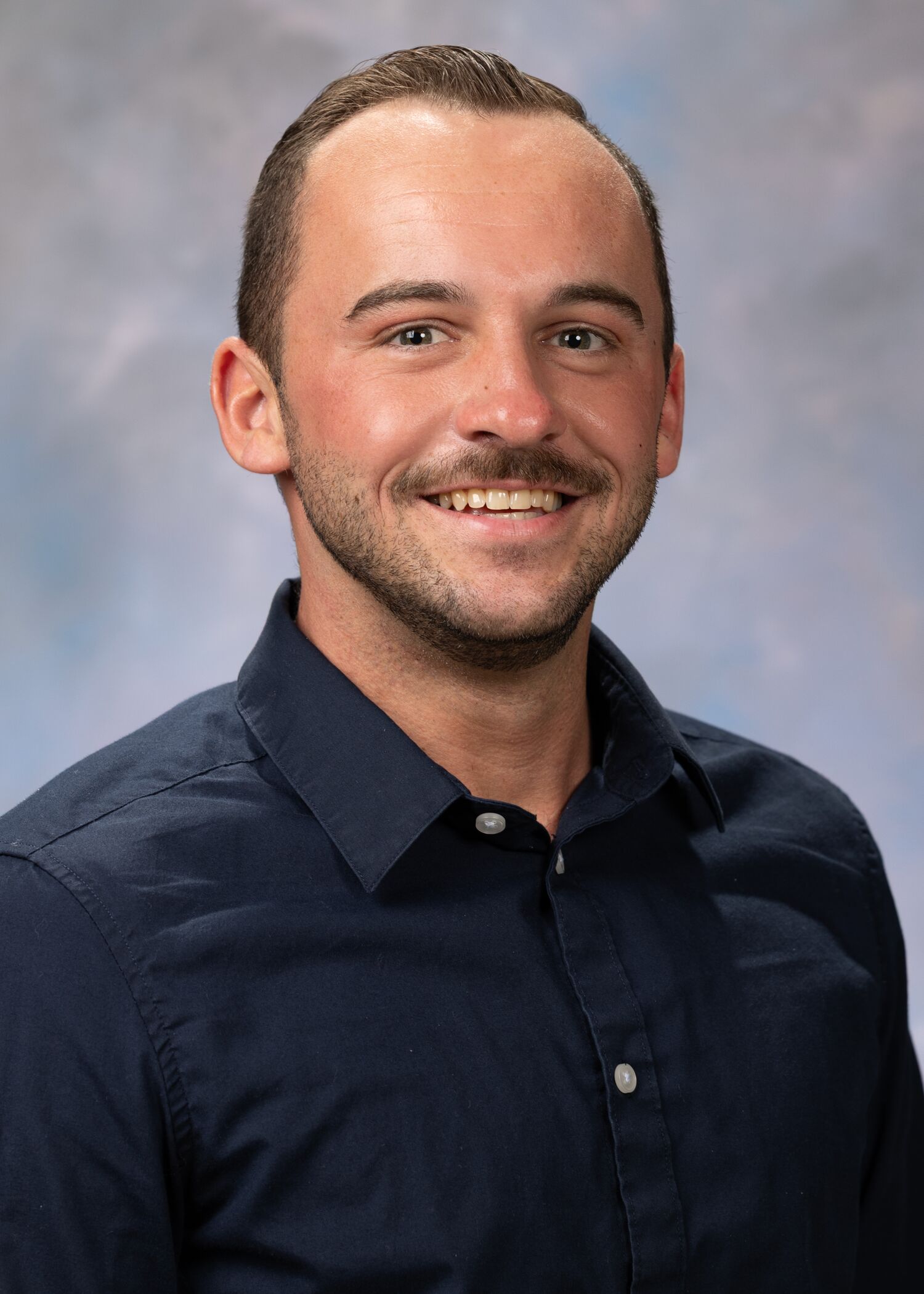 Portrait of Austin Guyton. Austin has short hair and facial hair. He is wearing a navy-blue button up shirt.
