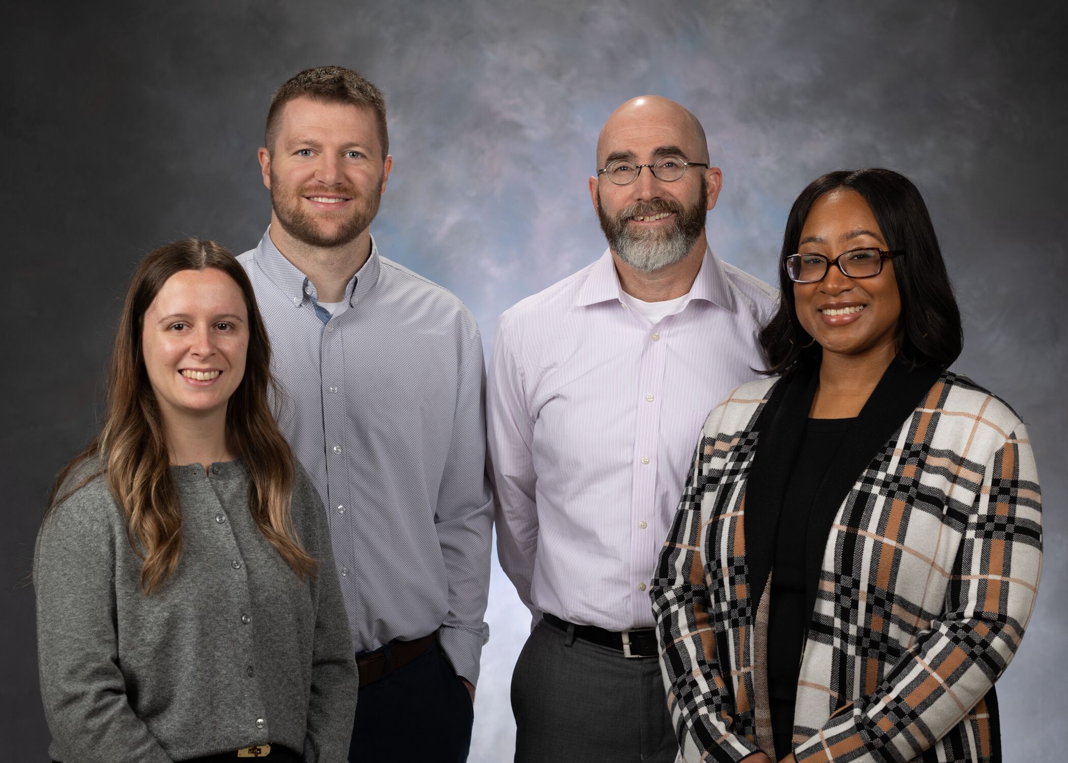 Four individuals in business casual attire posing for a group photo in front of a neutral, cloudy background. Two are standing in the front row and two in the back row.