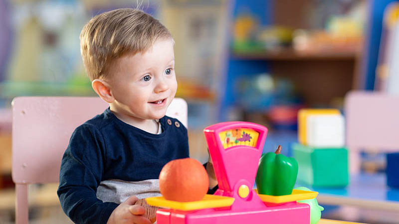 Happy toddler boy plays with a colorful toy food scale and plastic vegetables in a bright playroom.