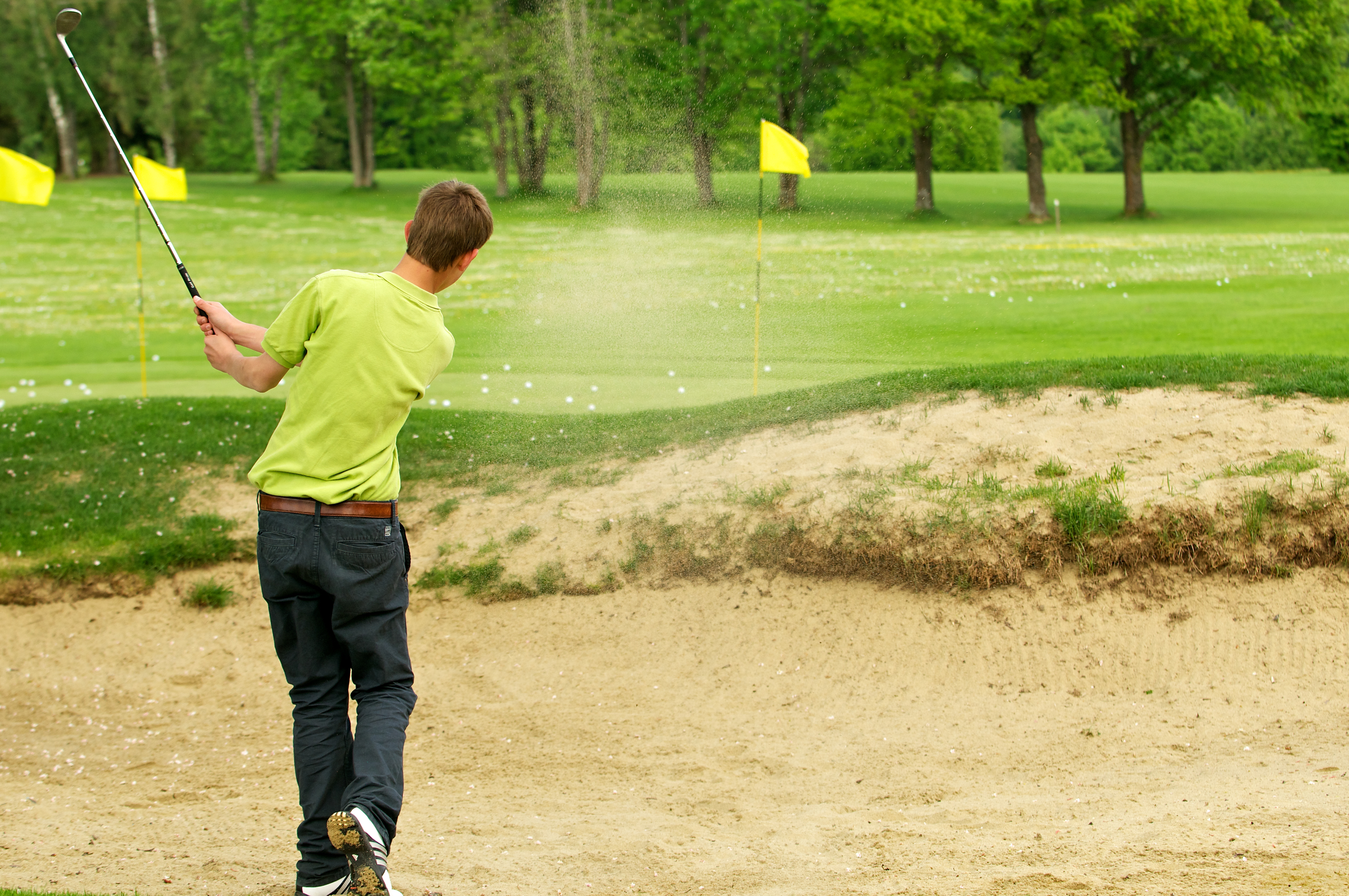 Person swinging a golf club from a sand bunker on a golf course, sending sand into the air. Several yellow flags and green grass are visible in the background with trees lining the course.